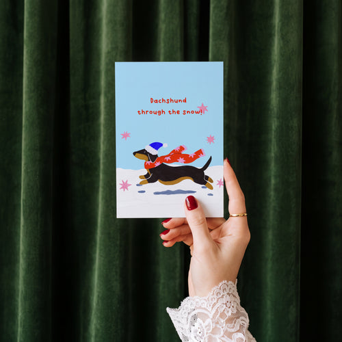 A hand with red nail polish and a white lace cuff holds a greeting card in front of a dark green velvet curtain. The card features an illustration of a black and tan Dachshund wearing a blue hat and a red scarf, with the text 'Dachshund through the snow!'.