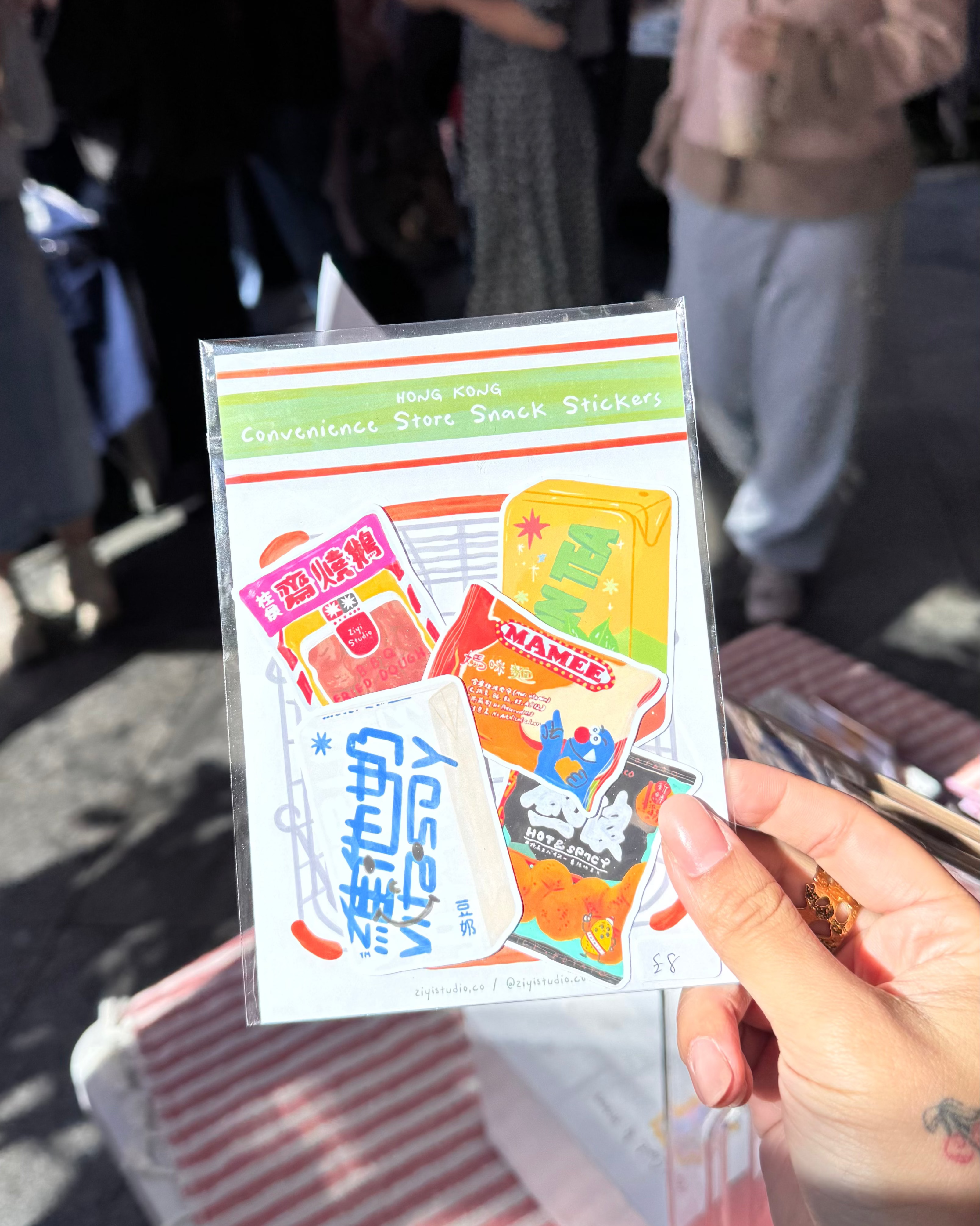 A hand holds a clear plastic sleeve containing a sheet of colorful retro-style snack and candy stickers, photographed at an outdoor market with blurred people in the background.