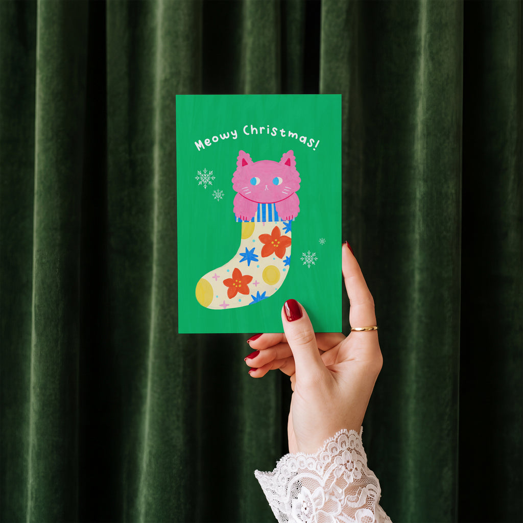 Meowy Christmas card held in hand against a green velvet curtain backdrop