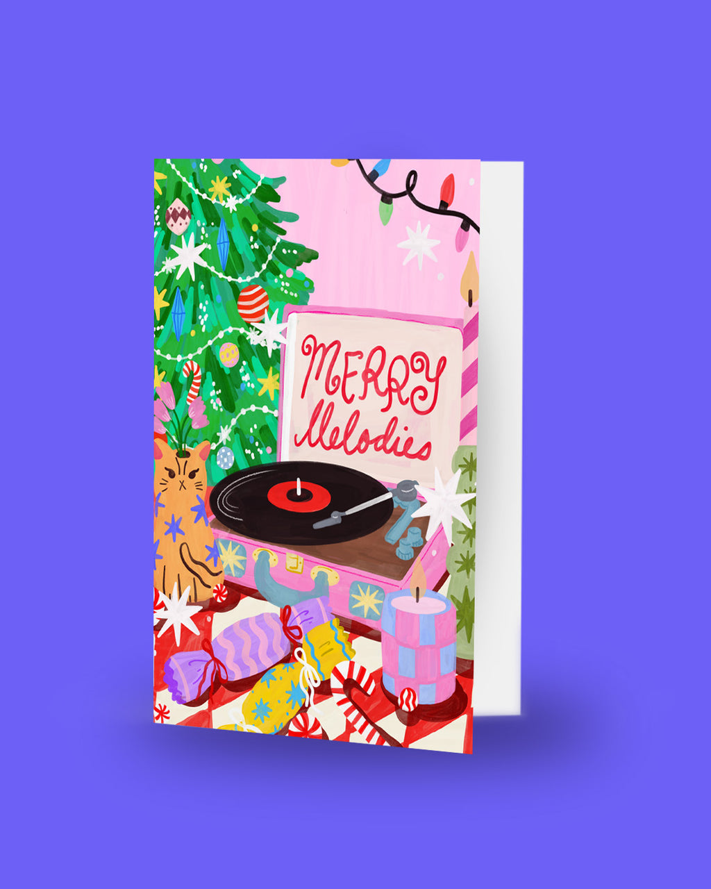 A festive Christmas greeting card featuring a pink background with a decorated tree, string lights, wrapped gifts, and a turntable with the words 'Merry Melodies' in red script. The card is shown standing upright against a solid purple background.
