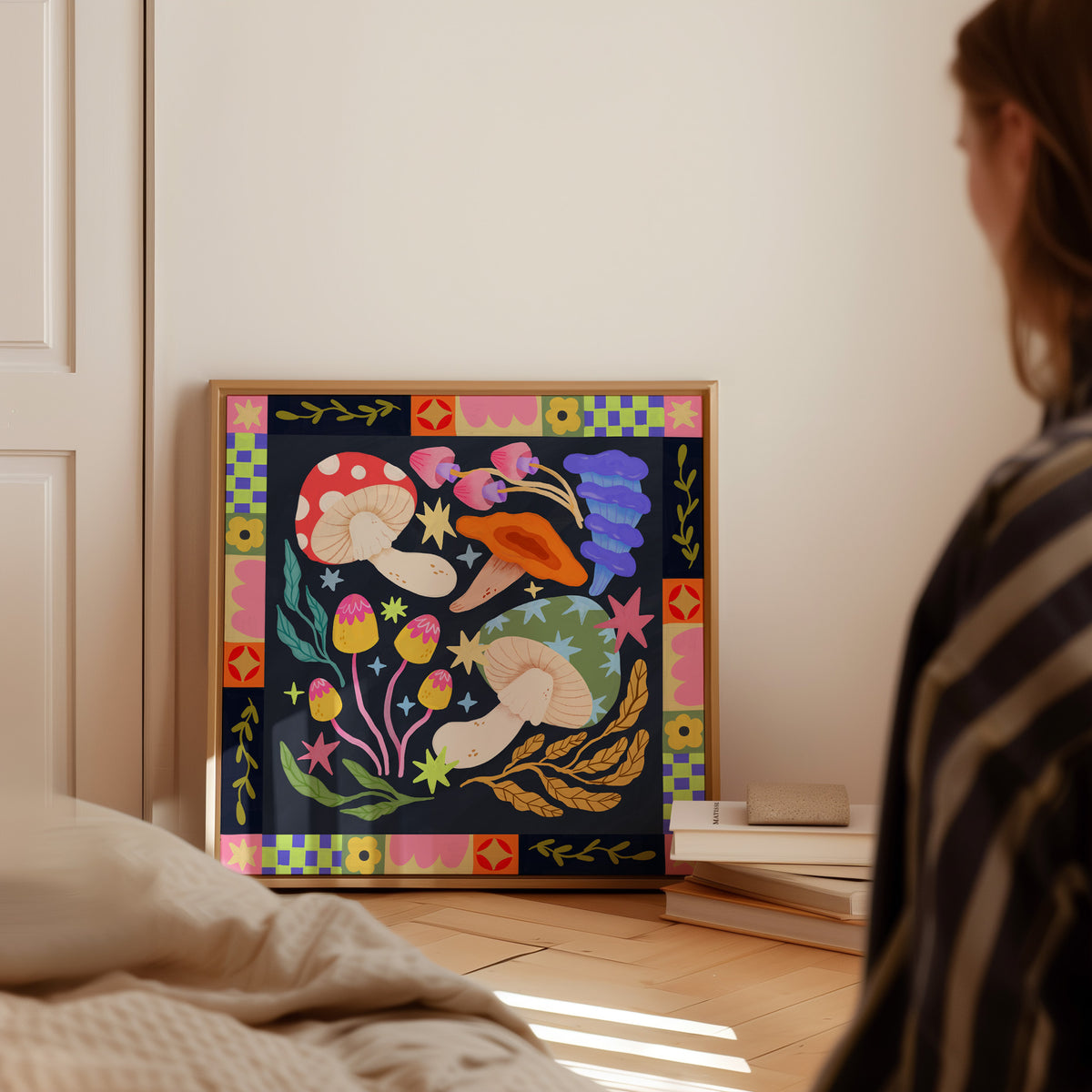 A framed colorful folk-art style print featuring mushrooms, flowers, and geometric borders leaning against a wall in a bedroom, with part of a person visible on the right.