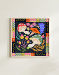A framed square art print featuring a whimsical folk-art style illustration of mushrooms, flowers, stars, and leaves on a dark background, bordered by a colorful geometric pattern. The framed print is mounted on a plain off-white wall.