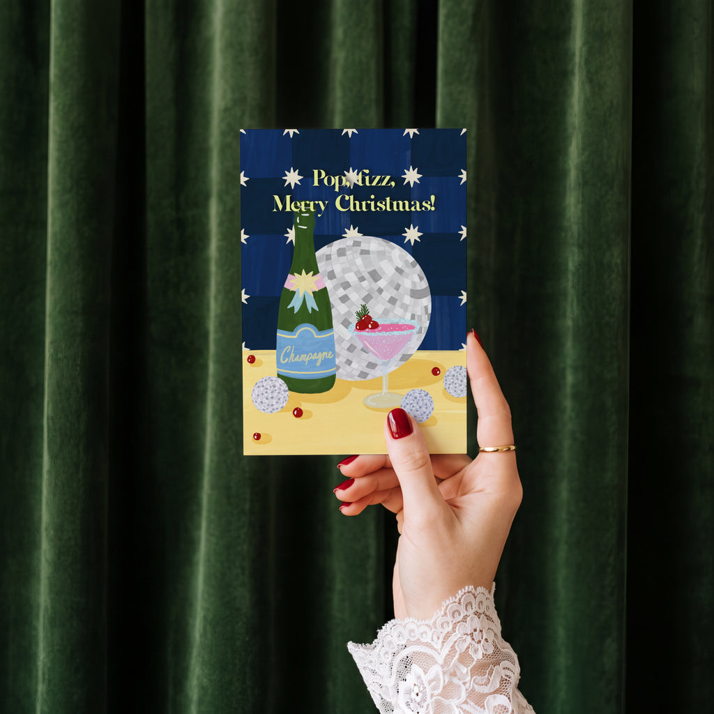 Pop Fizz Merry Christmas card held in hand against a green velvet curtain backdrop