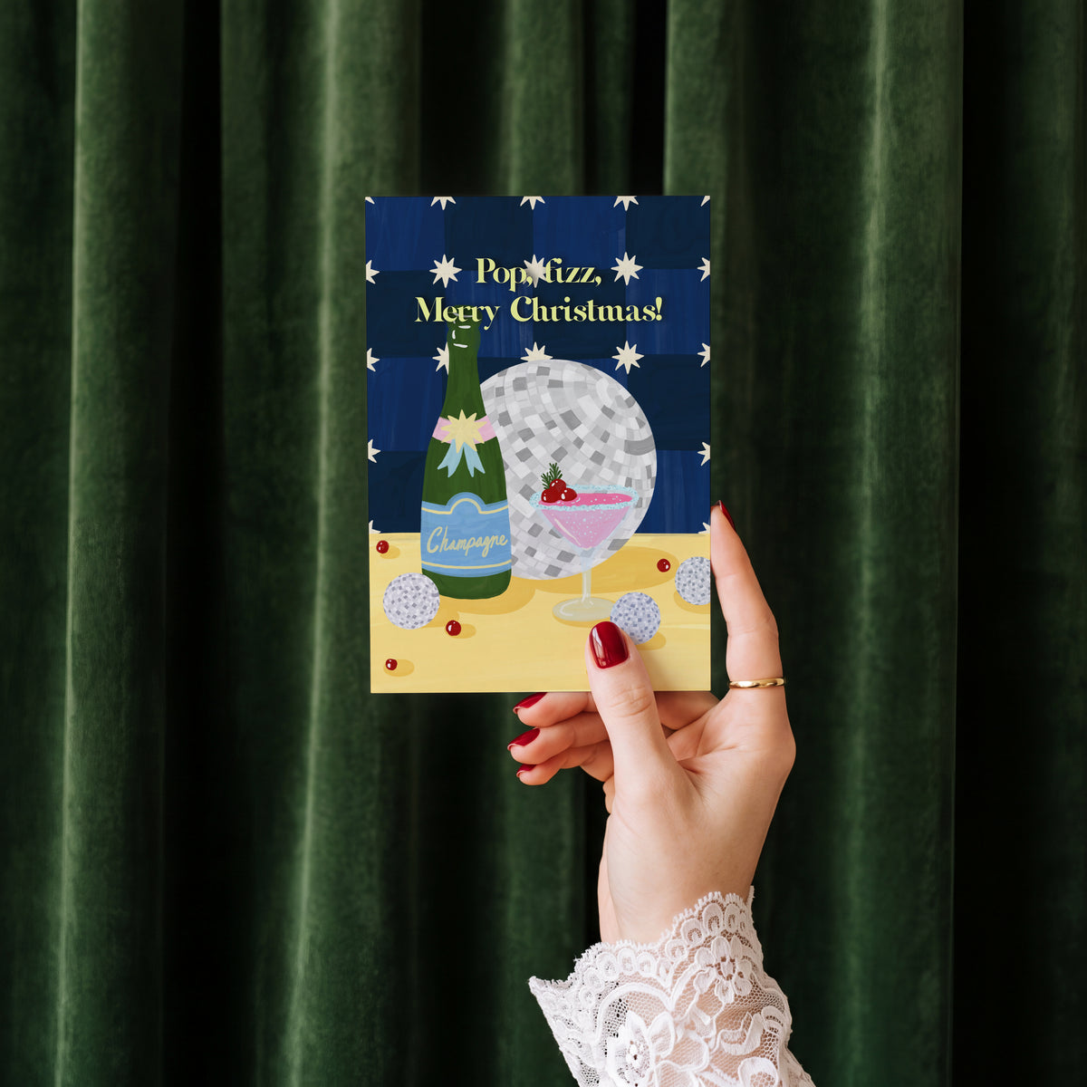 Pop Fizz Merry Christmas card held in hand against a green velvet curtain backdrop