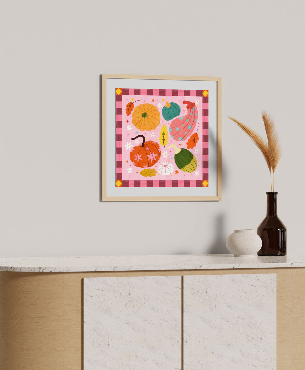 A framed art print featuring colorful autumn pumpkins and gourds on a pink gingham background, styled on a light gray wall above a wooden cabinet with a vase and dried grass.