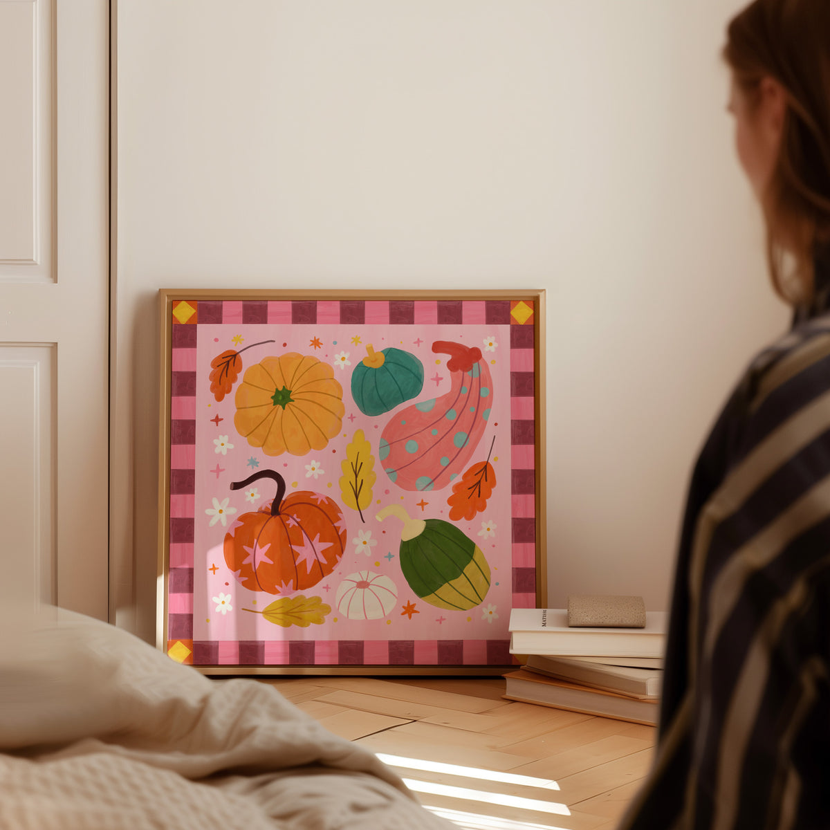 A framed autumn-themed art print featuring colorful pumpkins, acorns, and leaves on a pink gingham background, leaning against a wall in a softly lit bedroom. A person is partially visible in the foreground, looking at the artwork.