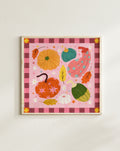 A framed autumn-themed art print featuring colorful pumpkins, gourds, leaves, and stars on a pink background with a dark pink gingham border, hung on a light beige wall.