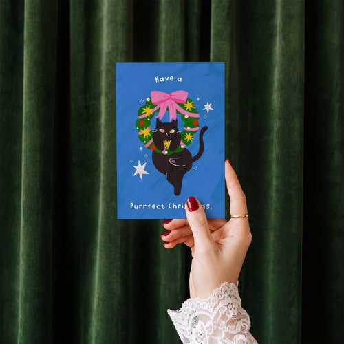 Have a Purrfect Christmas card held in hand against a green velvet curtain backdrop