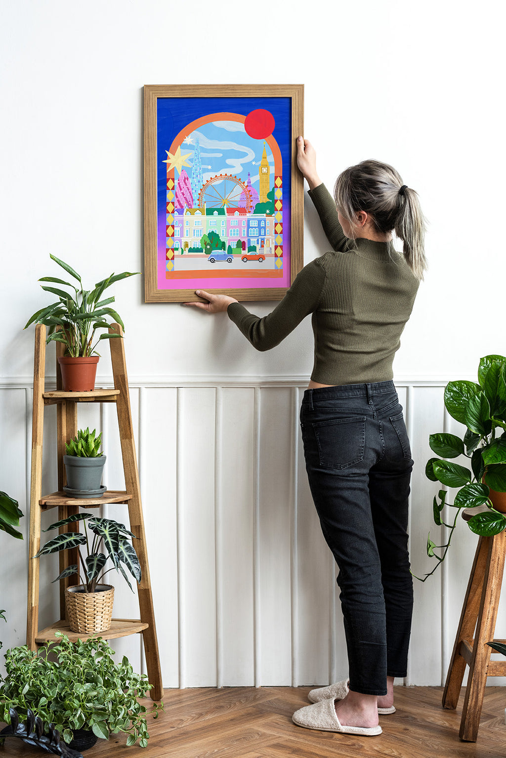 Person hanging a colorful framed artwork on a wall with plants around