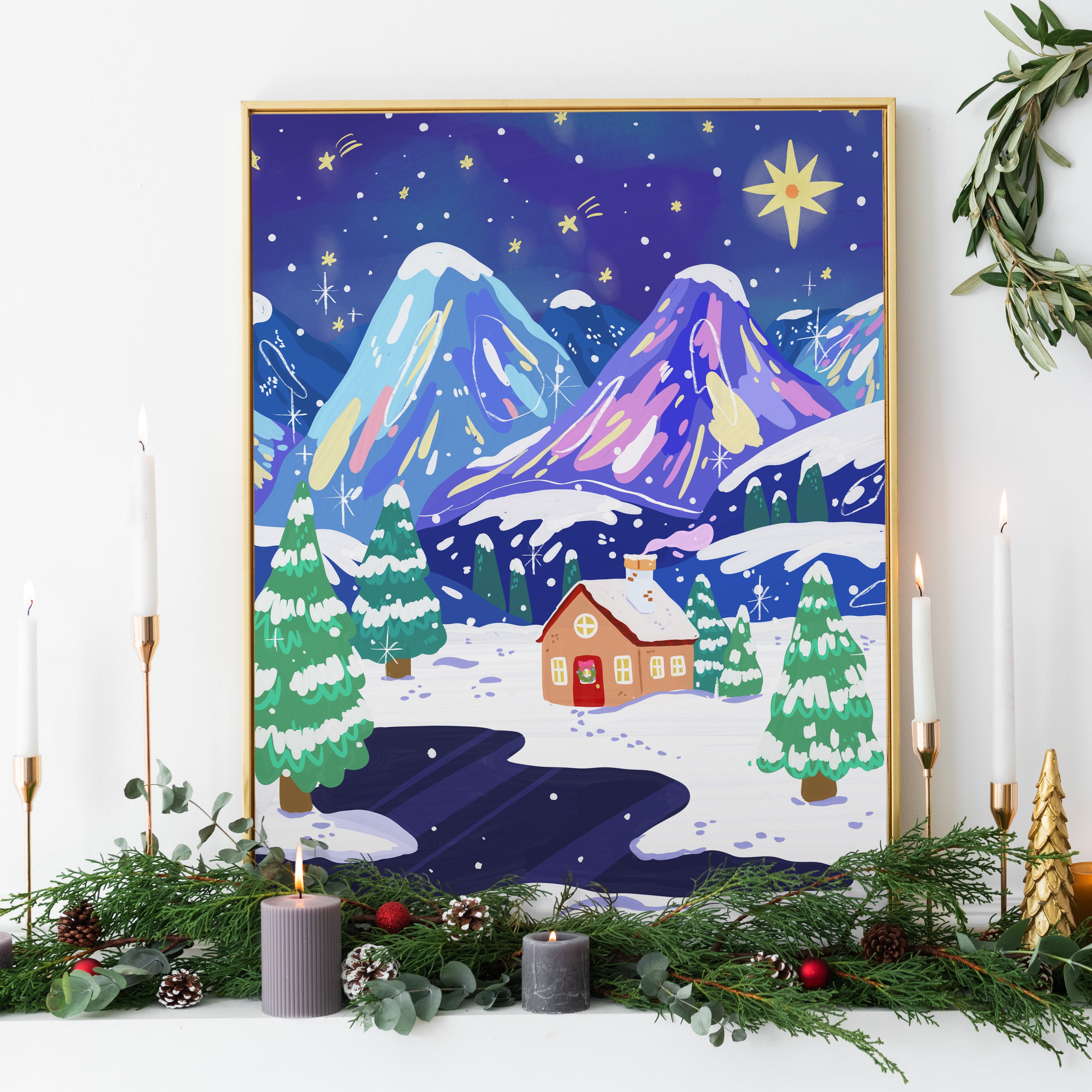 Framed winter landscape painting with a house and mountains, surrounded by candles and greenery.
