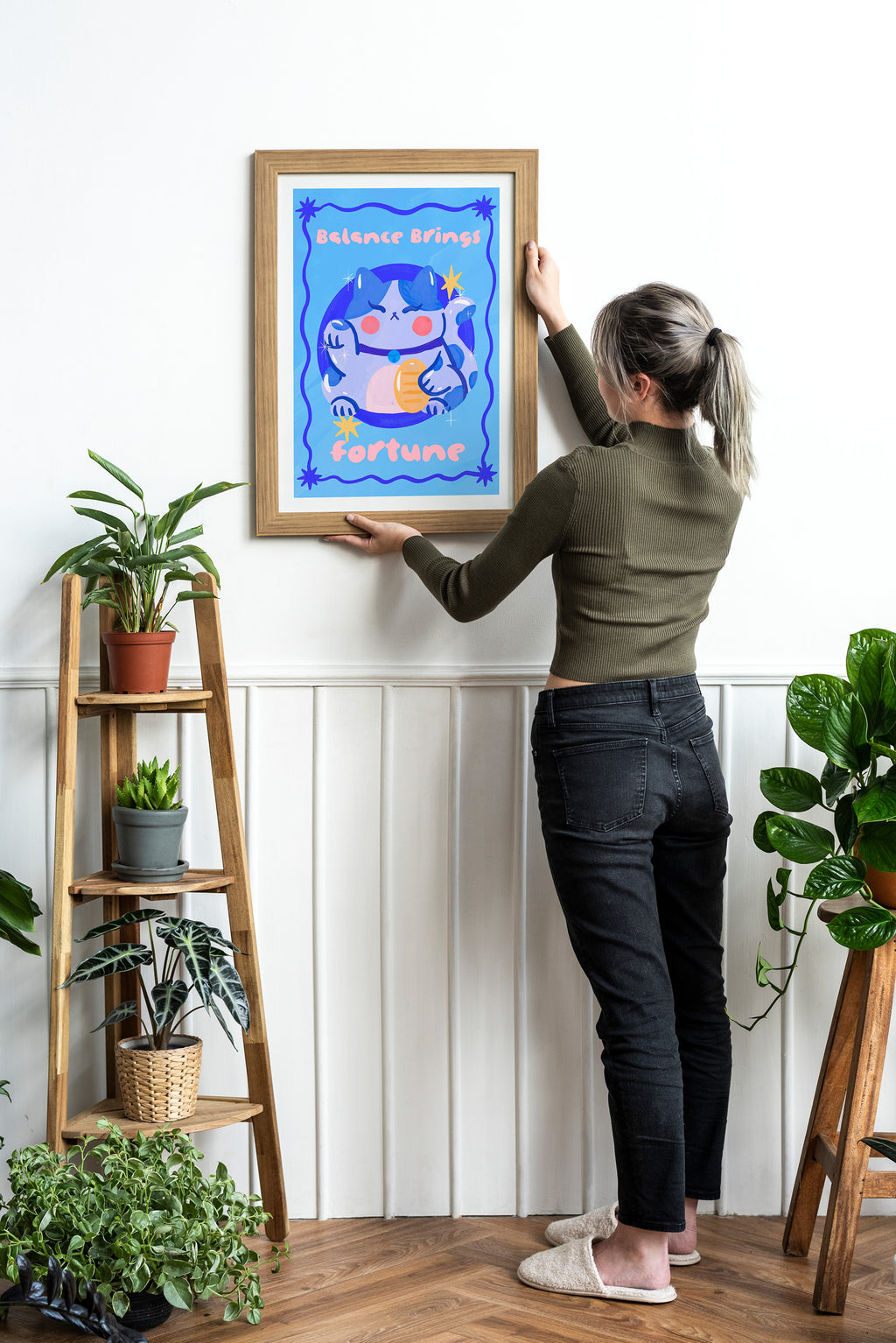 Person hanging a framed artwork on a wall with plants around