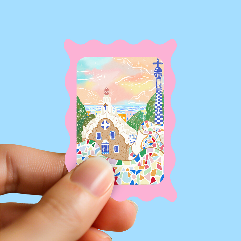 Mini hand holding a pastel pink stamp-style Barcelona sticker featuring Park Güell mosaic tiles and whimsical architecture, against a light blue background.