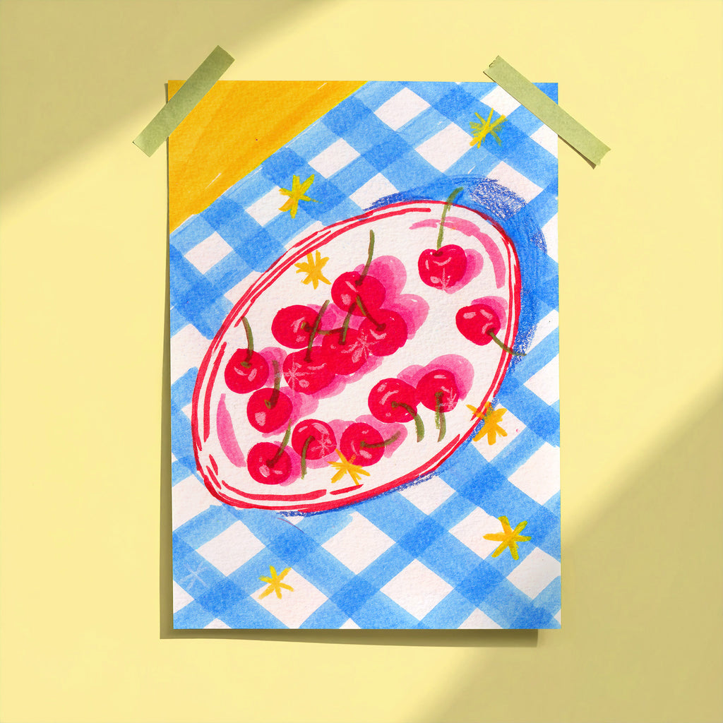 Bowl of Cherries art print taped to yellow wall showing red cherries on oval plate with blue gingham background, limited edition print by Ziyi Studio London
