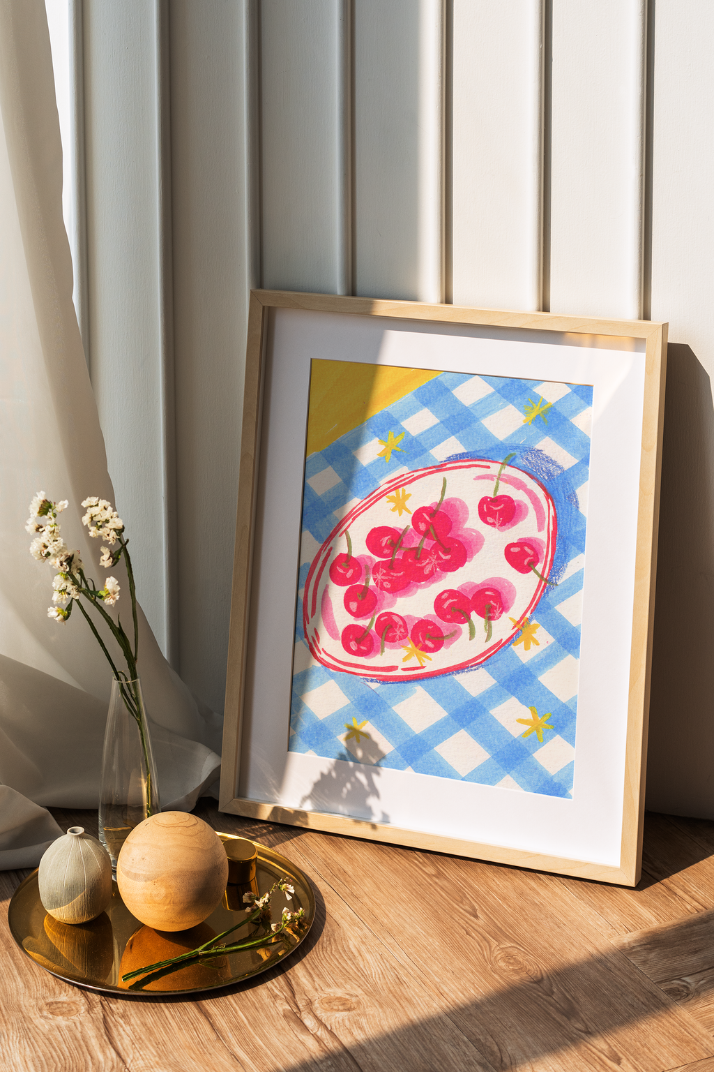 A framed art print featuring a watercolor illustration of cherries on a blue and white checkered plate, leaning against a paneled wall on a wooden floor next to decorative vases.