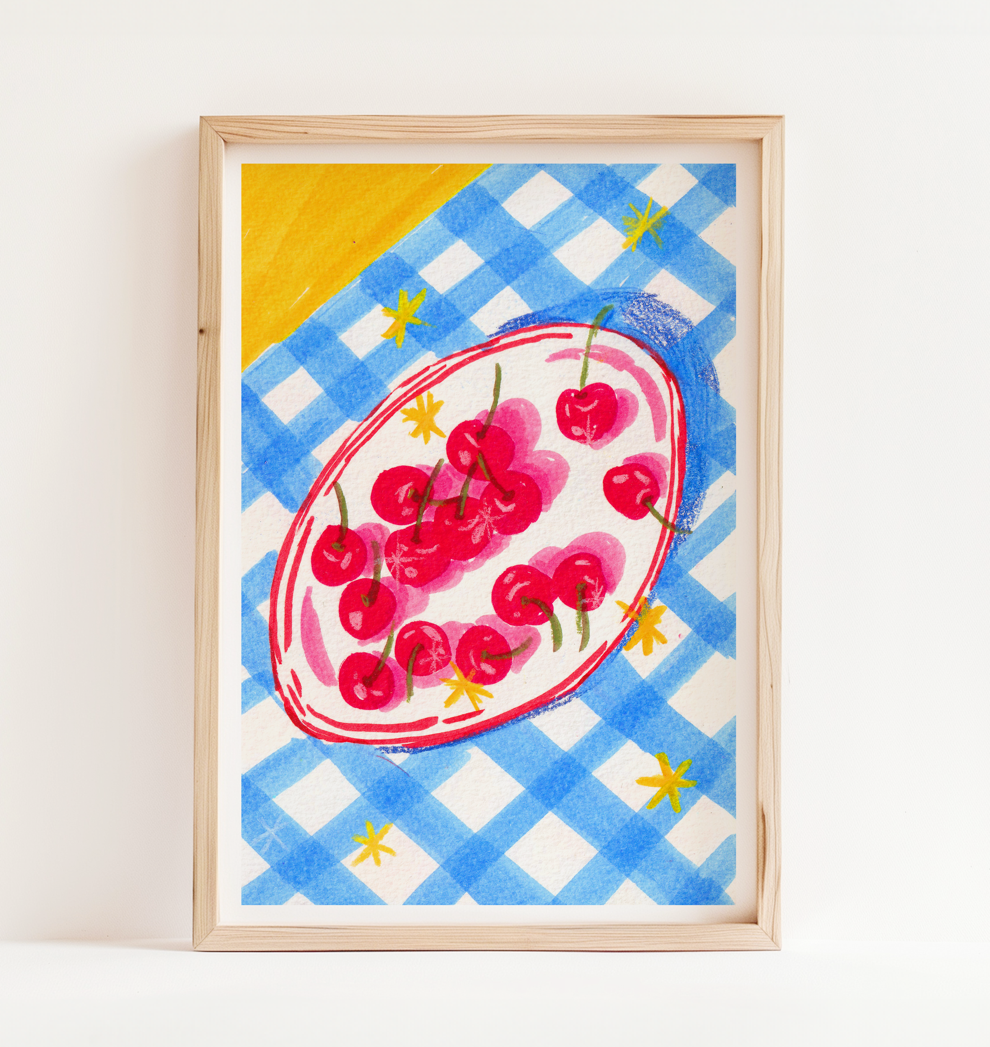 A framed art print featuring a whimsical illustration of a plate of cherries on a blue and white checkered background with yellow accents, set in a light natural wood frame against a white wall.