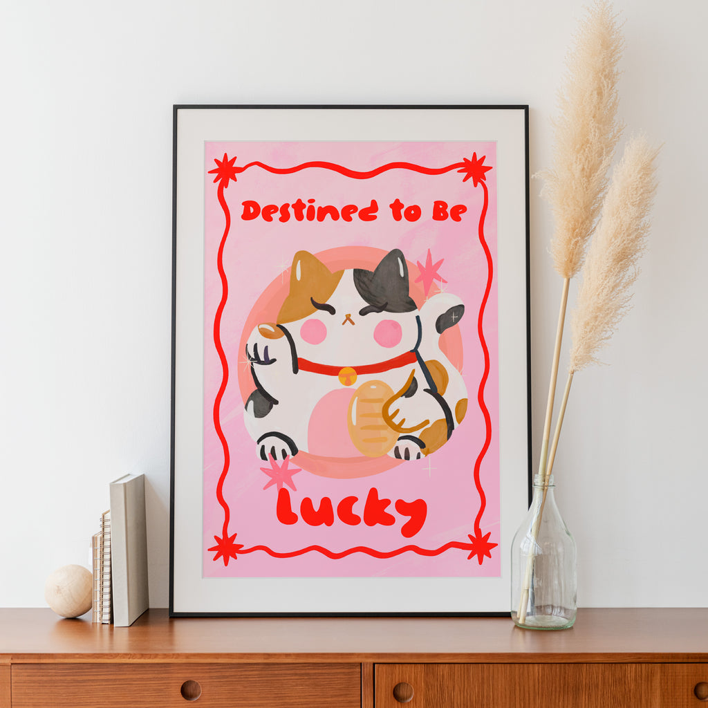 Framed artwork with a cat illustration and text 'Destined to be lucky' on a pink background, displayed on a wooden surface.