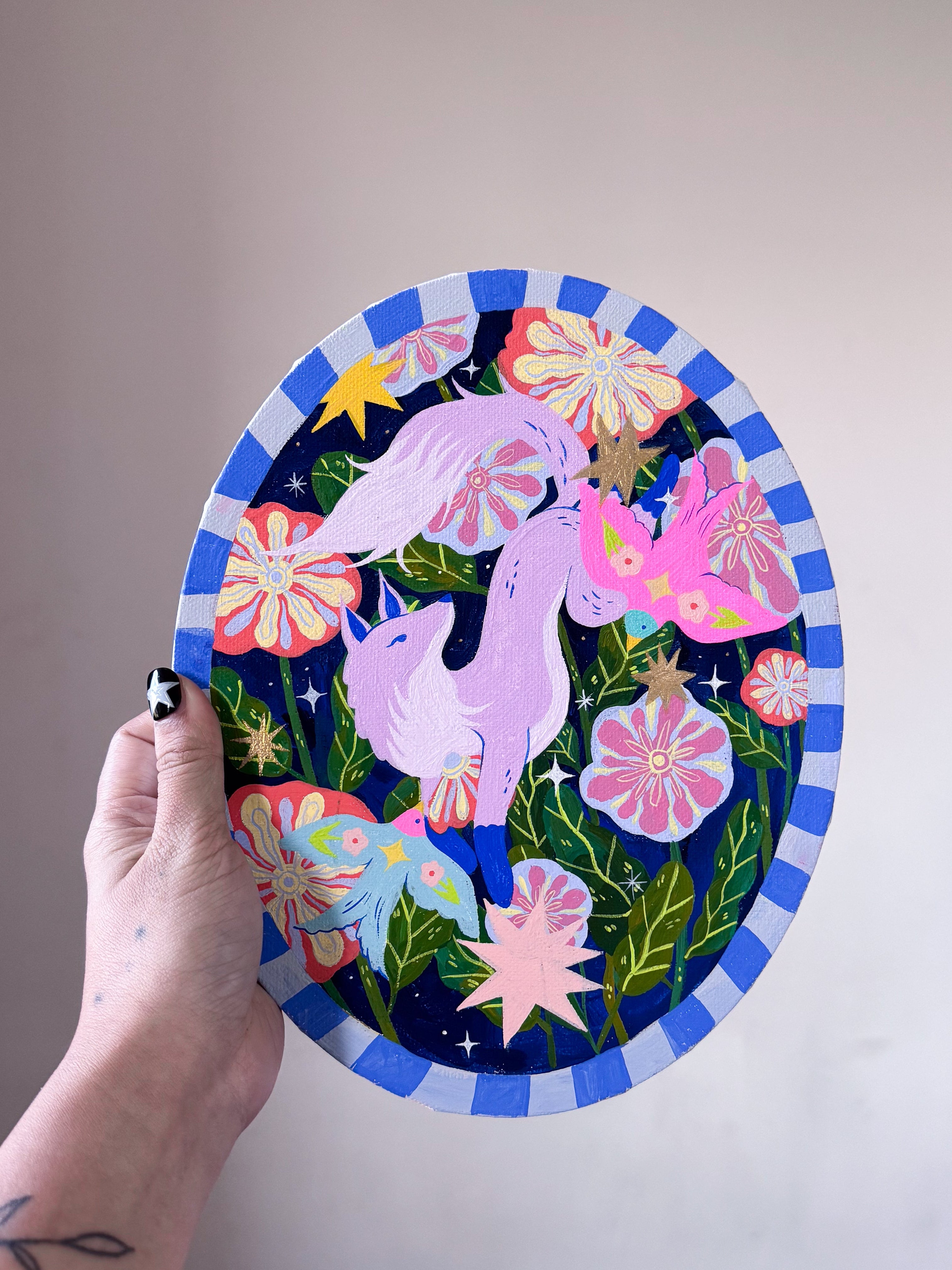 Hand holding a colorful paper cutout of a unicorn with flowers against a plain wall.