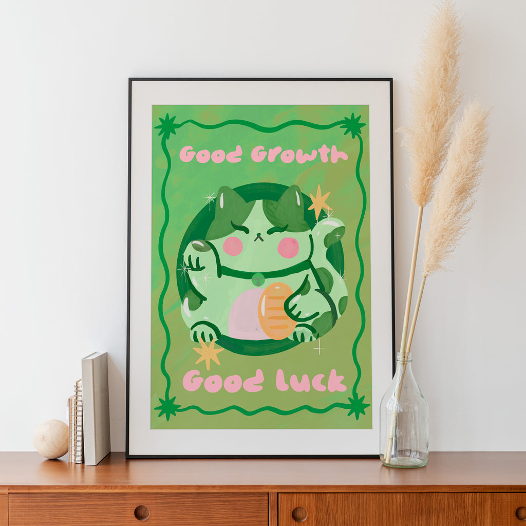 Framed illustration of a green creature with 'Good Growth' and 'Good Luck' text on a green background, placed on a wooden surface.