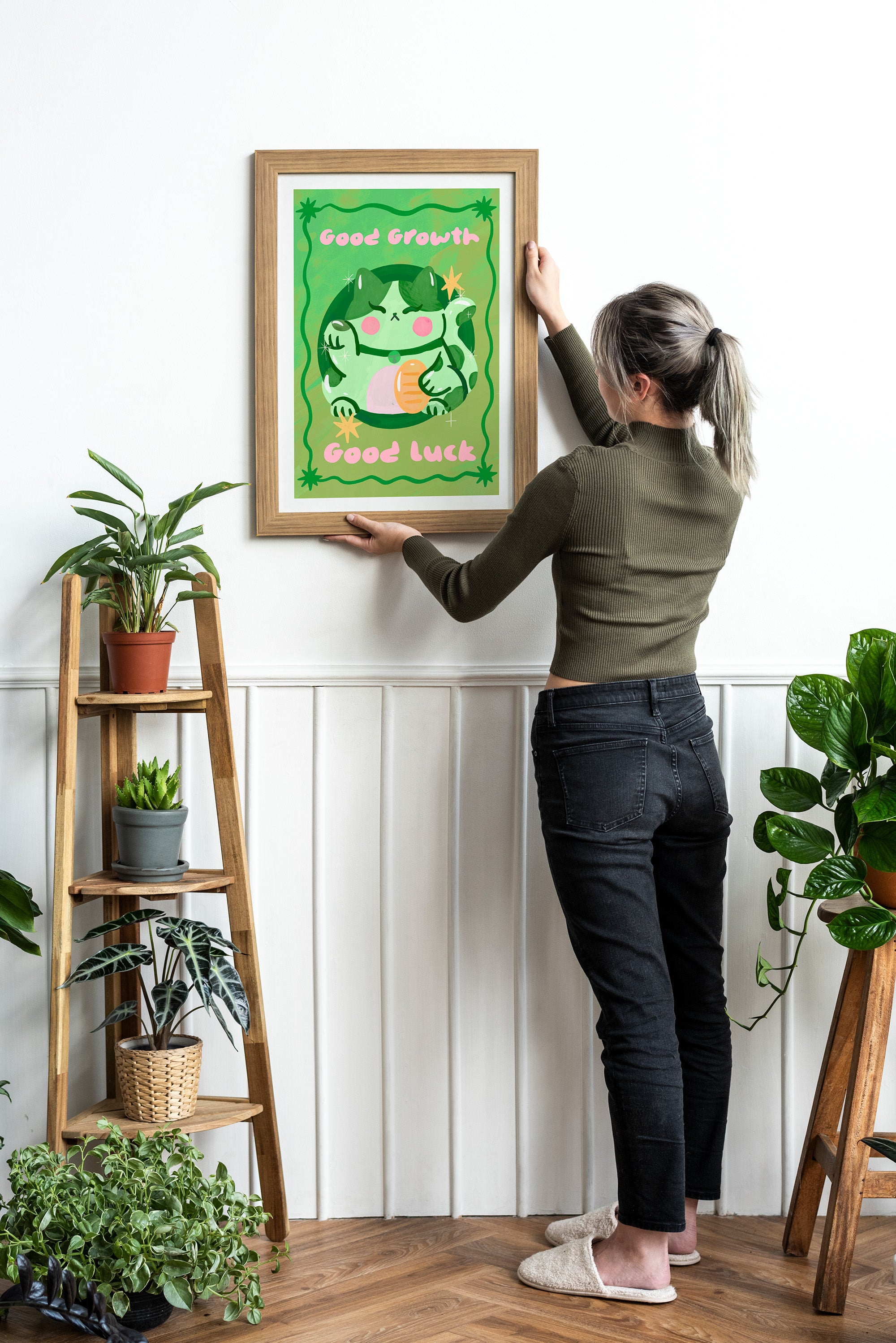 Person hanging a framed artwork of a cat on a wall with plants around