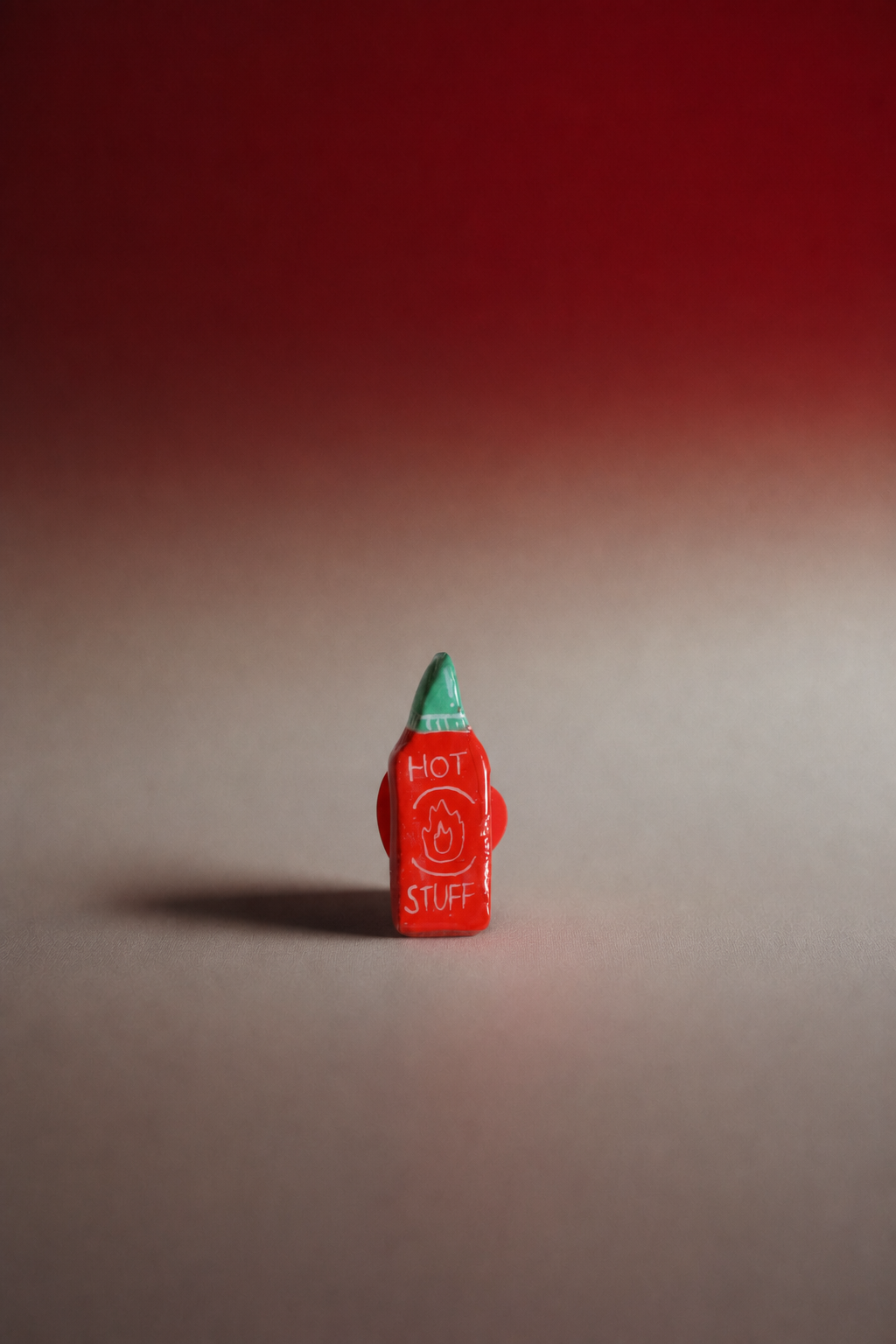 A small red novelty object shaped like a hot sauce bottle with a green pointed top, standing upright on a neutral surface against a red-to-gray gradient background. The front has white text reading 'HOT STUFF' with a small smiley face graphic.