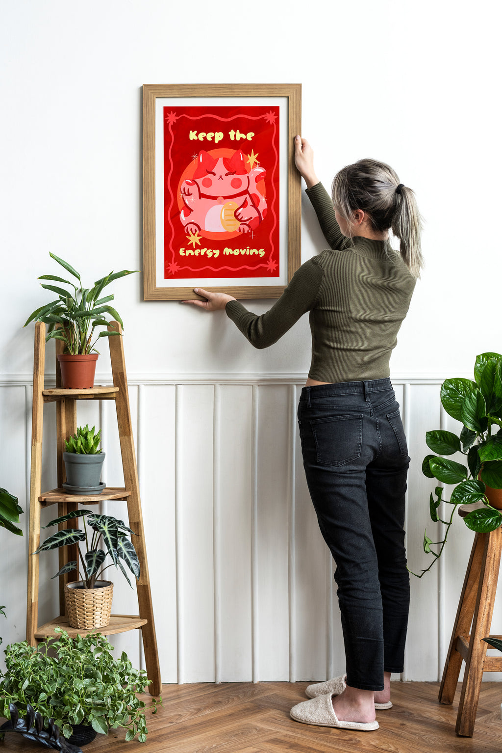 Person hanging a framed red poster on a wall with plants around