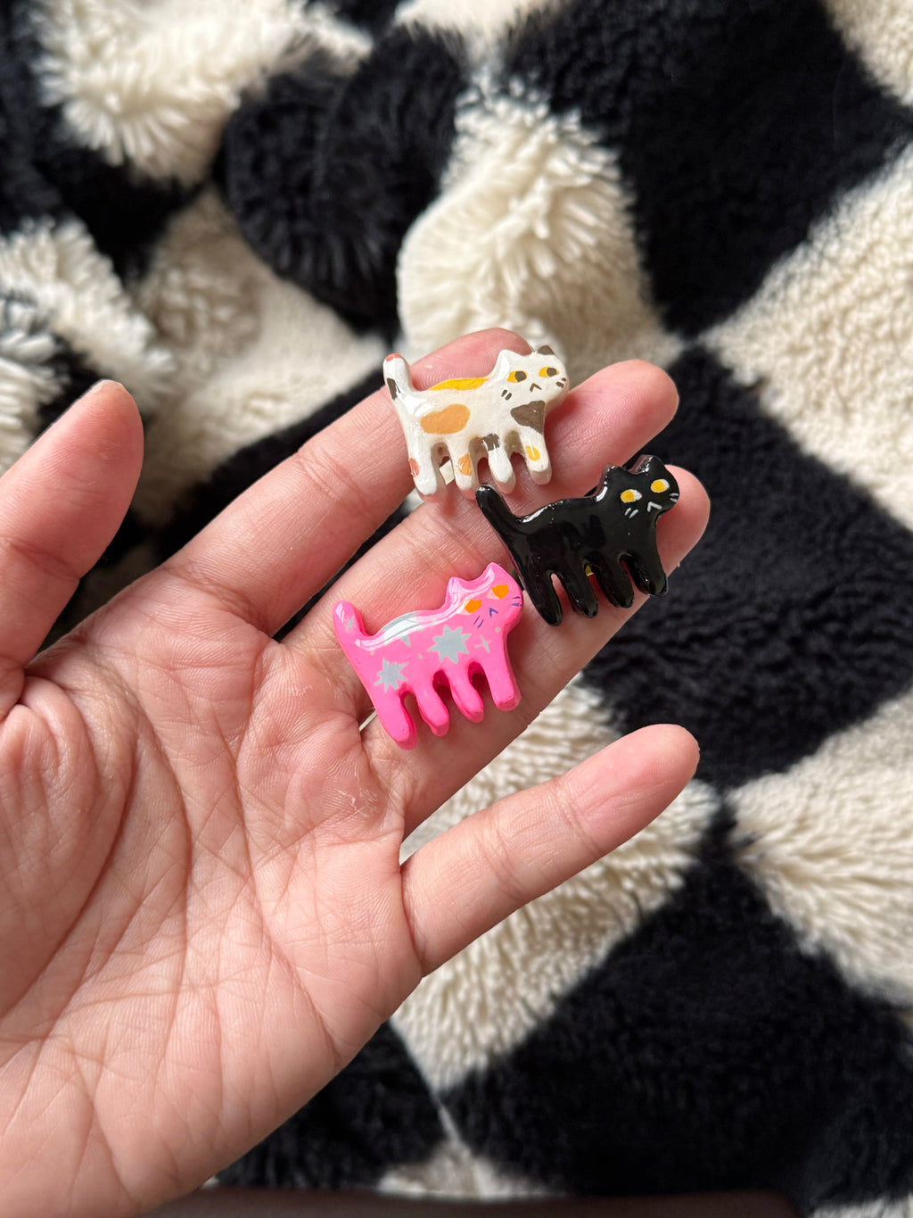 Hand holding two hair clips, one pink cat-shaped and one black cow-shaped, against a black and white patterned background.