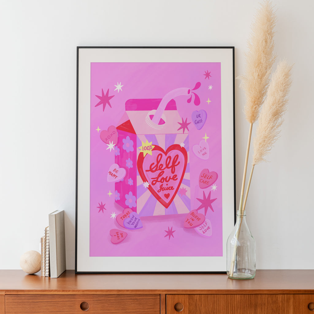 Framed artwork with 'Self Love Juice' design on a pink background, placed on a wooden surface.