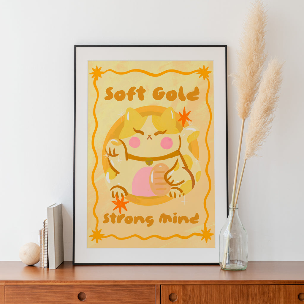 Framed artwork with a cartoon character and text 'Soft Gold Strong Mind' on a white wall.