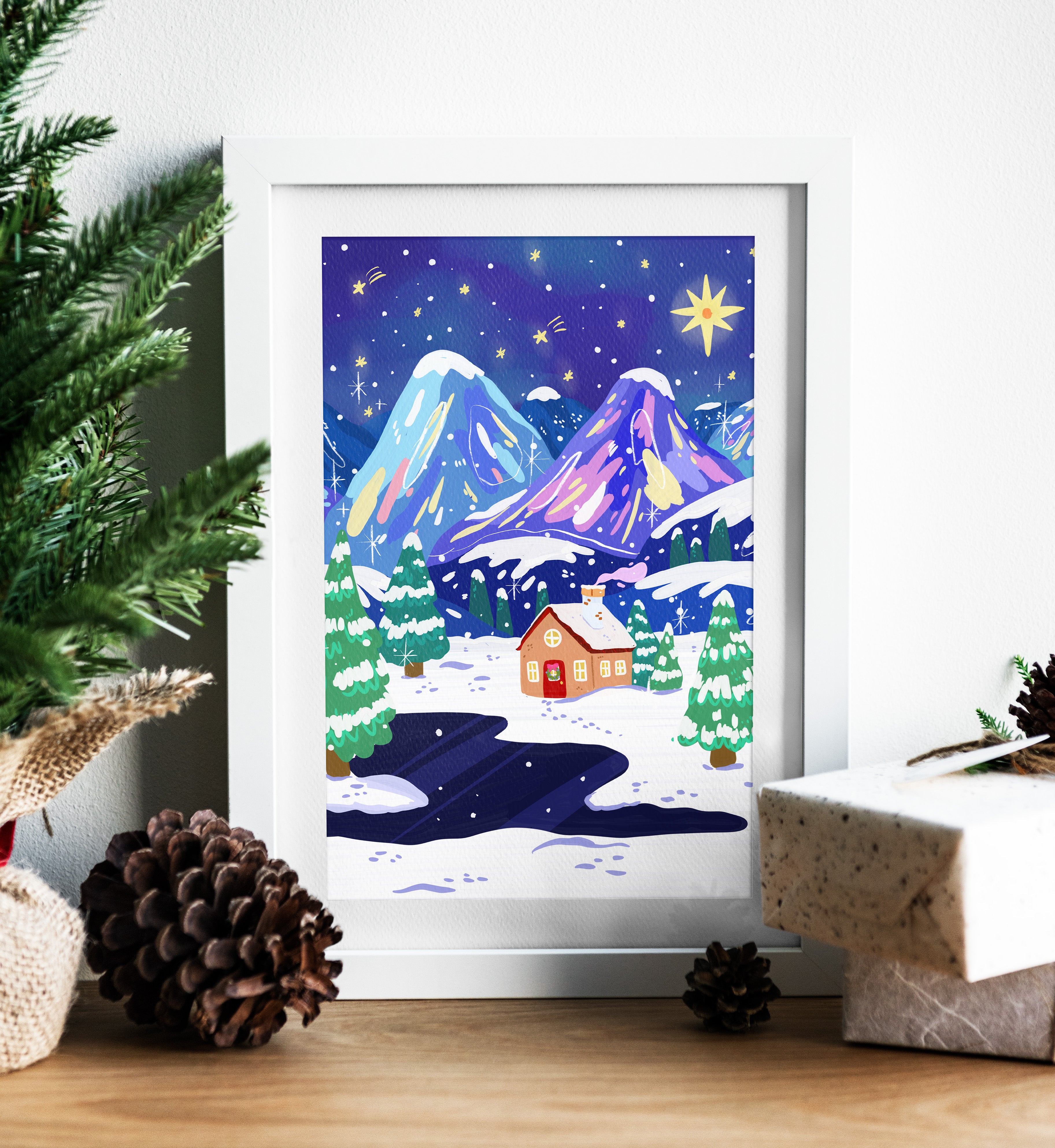 Framed winter scene print with mountains and a house, surrounded by Christmas decor.