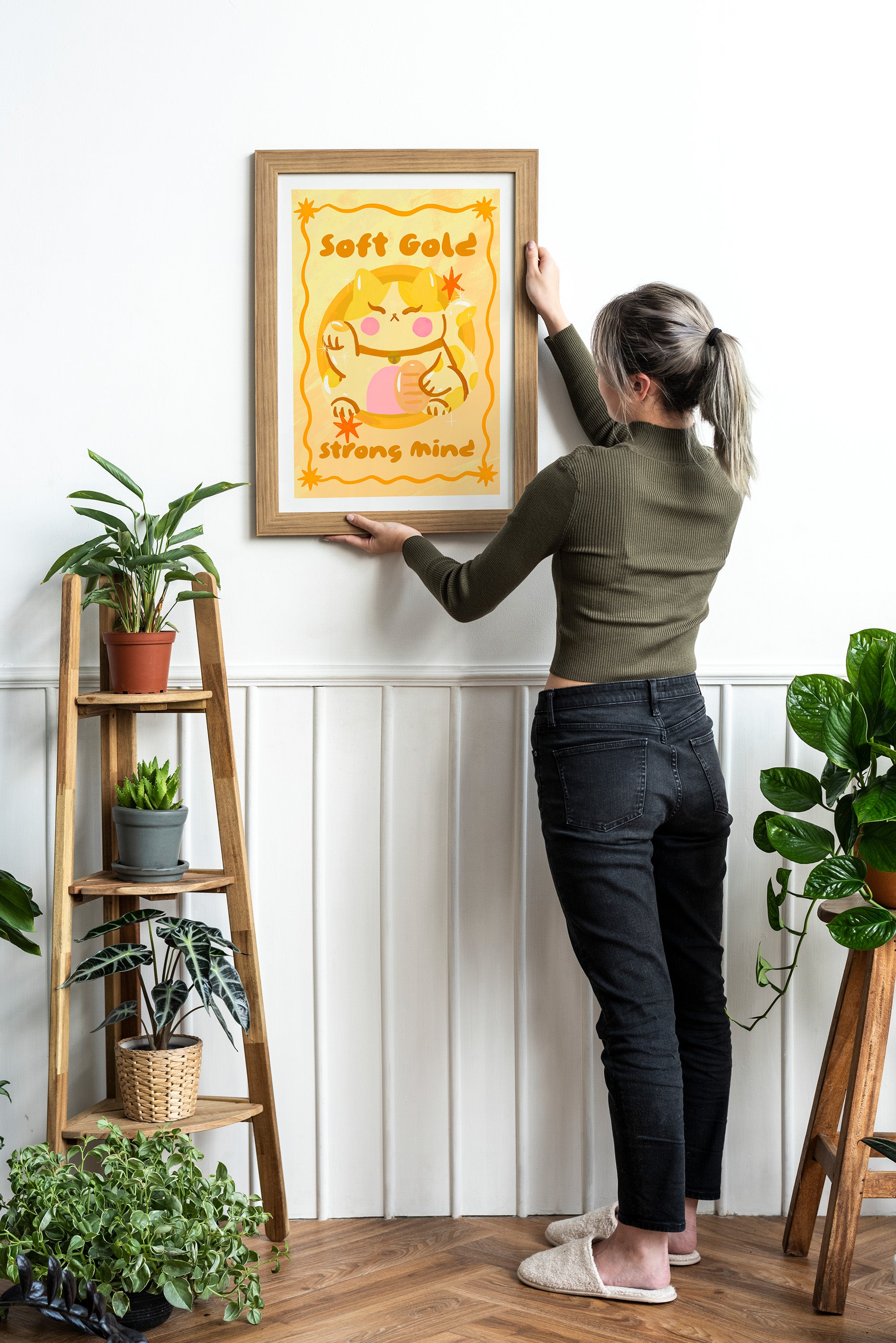 Person hanging a framed artwork on a wall with plants around